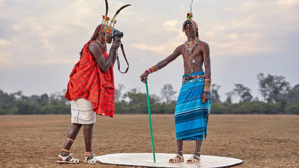 A Maasai elder in a red shuka stands beside a corrugated-iron radio shack on the edge of a Tanzanian trading town, holding a handheld shortwave receiver at dusk.