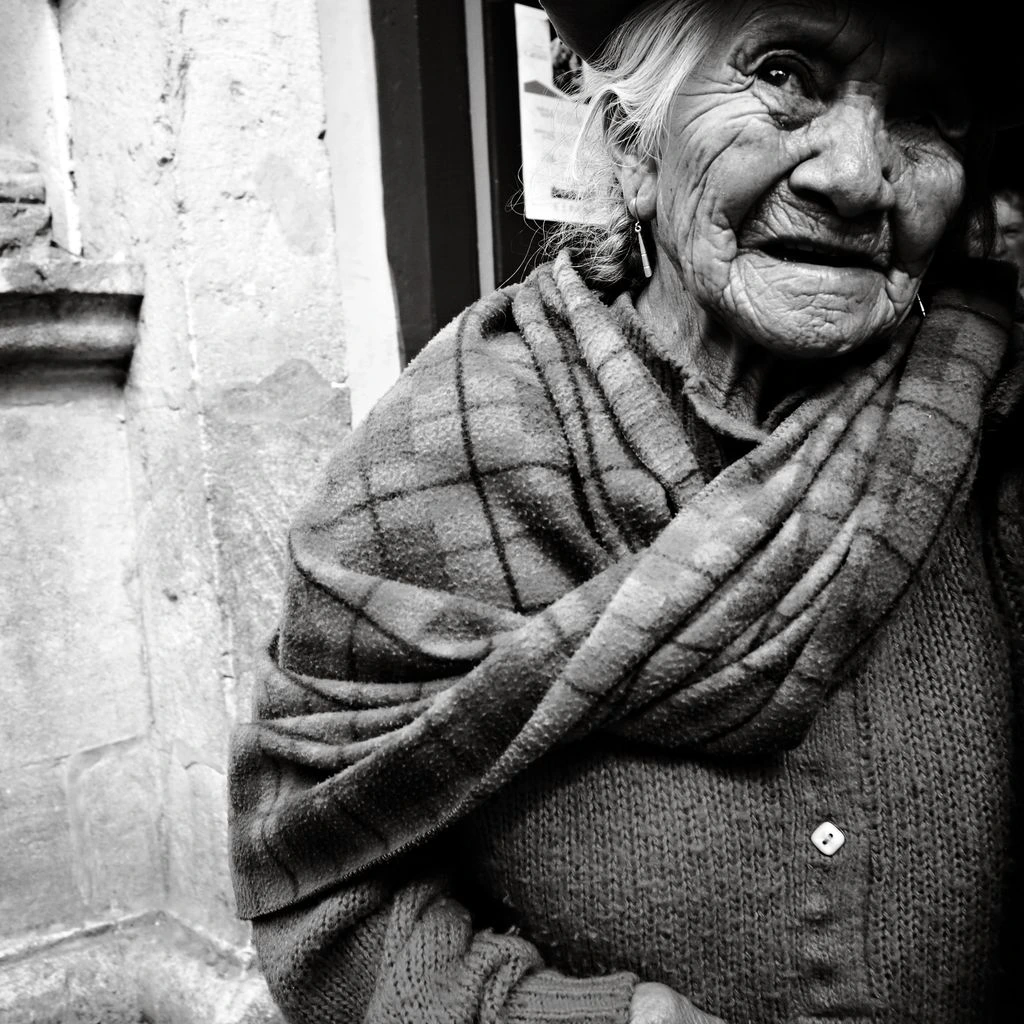 An elderly Candomblé priestess in white lace sits on a wooden veranda in Salvador, Bahia, speaking into a small handheld recorder held by an off-frame interviewer.